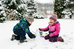 a picture of a toddler boy and girl dressed for winter with a winter jacket, hat, neck warmer, snow pants and boots and playing outside in the snow